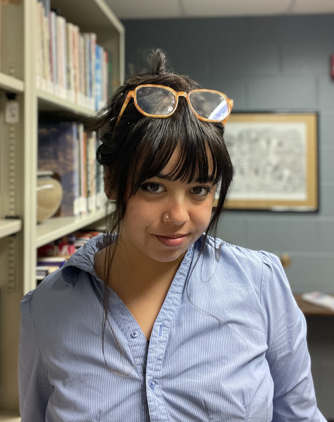 A young woman with dark hair, wearing glasses on her head, a nose ring, and a light blue button-up shirt, standing in front of bookshelves and a framed picture in a library or office.