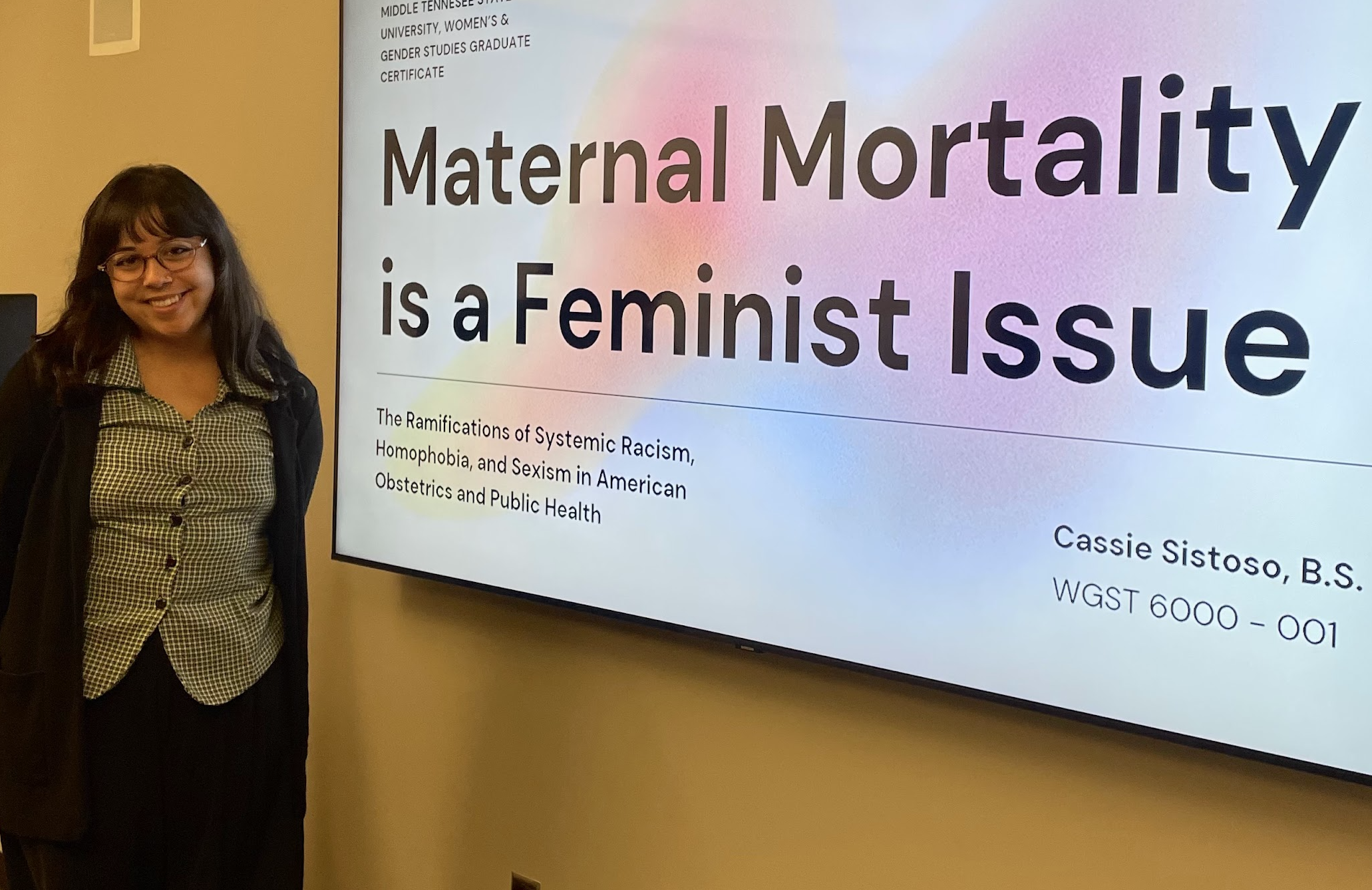 A woman with glasses and black hair standing next to a large presentation screen displaying a graduate certificate titled 'Maternal Mortality is a Feminist Issue.' She is wearing a checkered blouse and a dark cardigan, smiling at the camera.