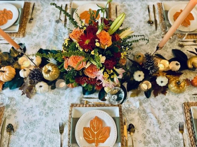 A tabletop decorated with a autumn themed centerpiece featuring flowers, pumpkins, gourds, pinecones, and candles. Placed around are plates with orange leaf designs, silverware, and napkins. Two tall candles are positioned on each side of the centerpiece.