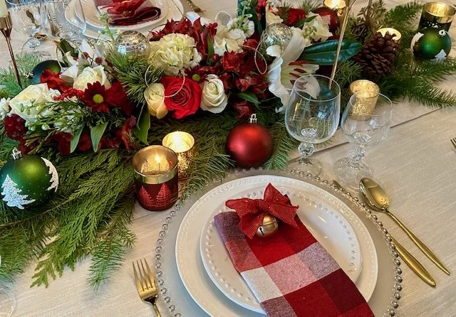 Christmas table setting with a white plate, red and plaid napkin, gold utensils, and decorative Christmas ornaments, candles, and greenery in the centerpiece.