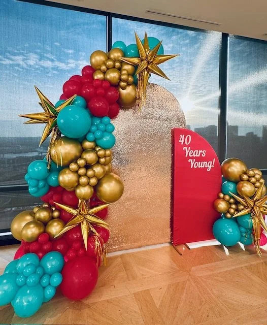 Decorative balloon arrangement with gold, red, and turquoise balloons, gold star-shaped balloons, and a red sign reading '40 Years Young!' in front of a glittering backdrop at an indoor celebration.