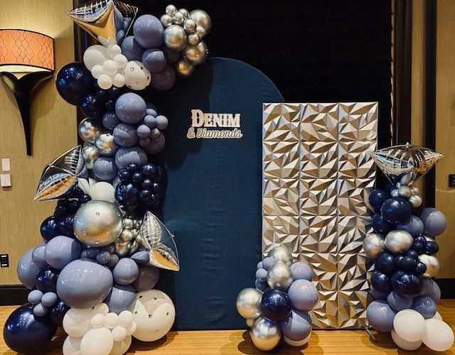 Decorative balloon arrangement with silver, white, blue, and purple balloons around a blue backdrop and a geometric gold panel, with a "DENIM & Diamond" sign.
