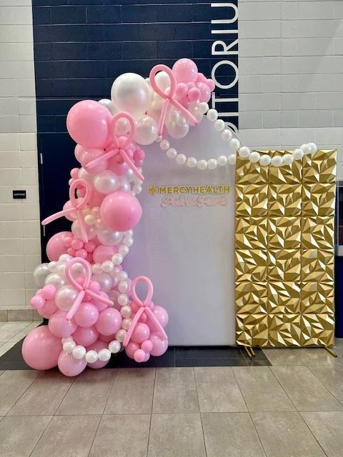 Pink and white balloon arrangement with bows, next to a gold geometric patterned partition, with a white backdrop displaying "#MERCYHEALTH".