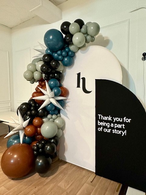Decorative balloon arch with various colors, including black, gray, teal, white, and brown, next to a white semi-circular backdrop with the letter 'h' and a black panel with a thank you message.