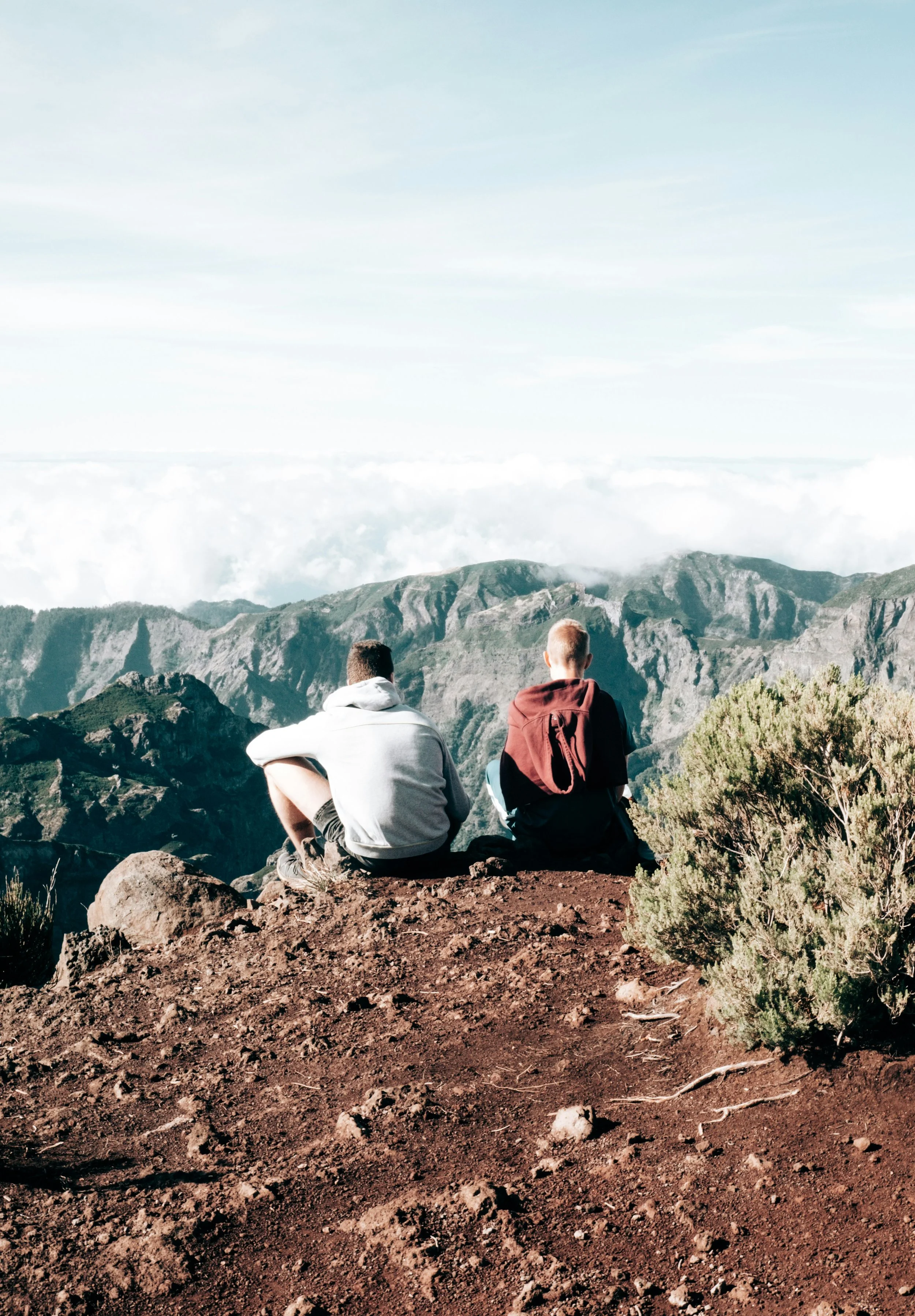 Two men sitting on a rocky cliff overlooking a mountain range and clouds, representing friendship and deep conversation.