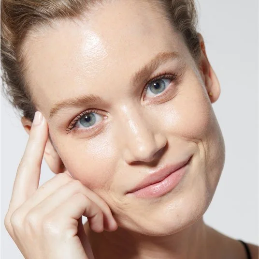 Close-up of a woman with blonde hair and blue eyes smiling gently, touching her temple with her right hand.