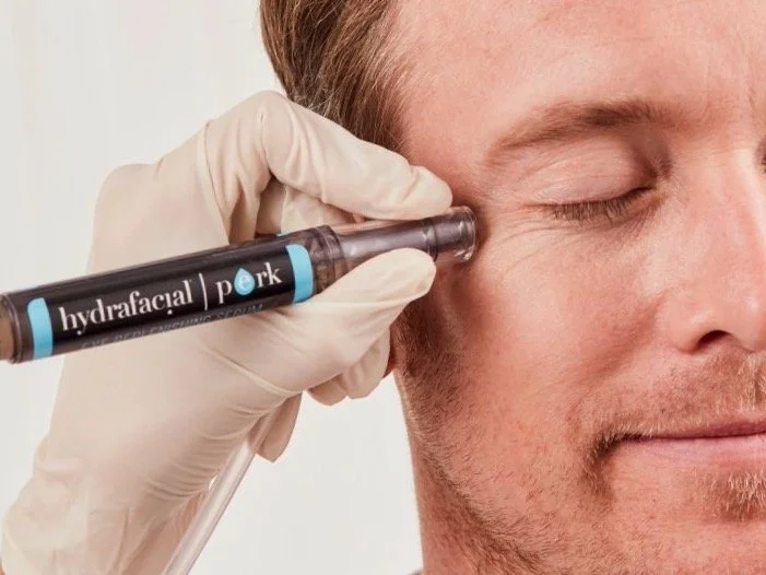 A person receiving a facial treatment with a syringe labeled 'hydrafacial' on their cheek, with eyes closed and wearing gloves.