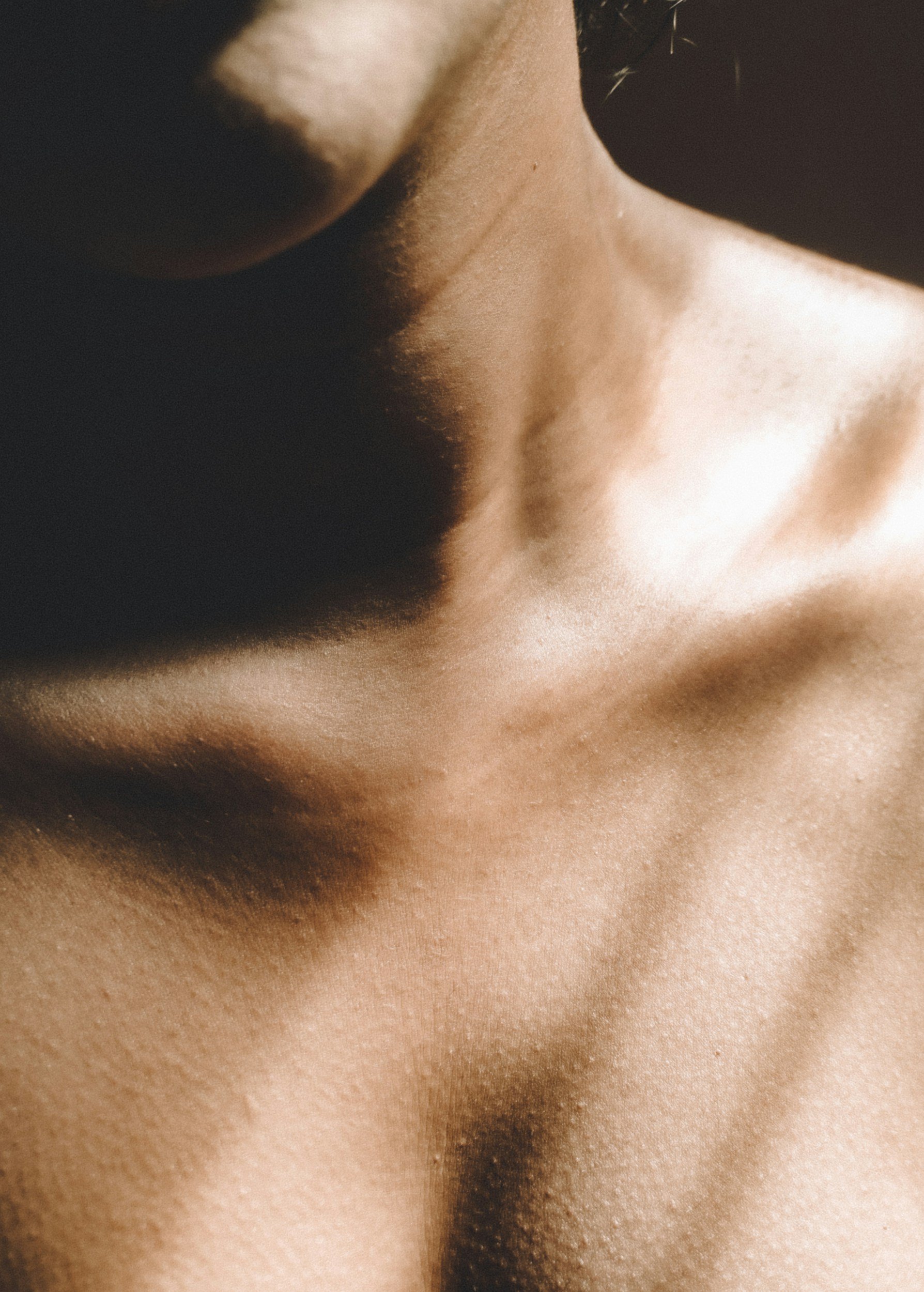 Close-up of a person's shoulder and collarbone, with smooth, tan skin and subtle lighting highlighting the texture and contours.