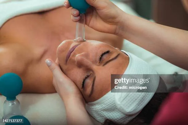 A woman receiving a facial treatment with a facial massager on her face, lying with eyes closed and wearing a headband.