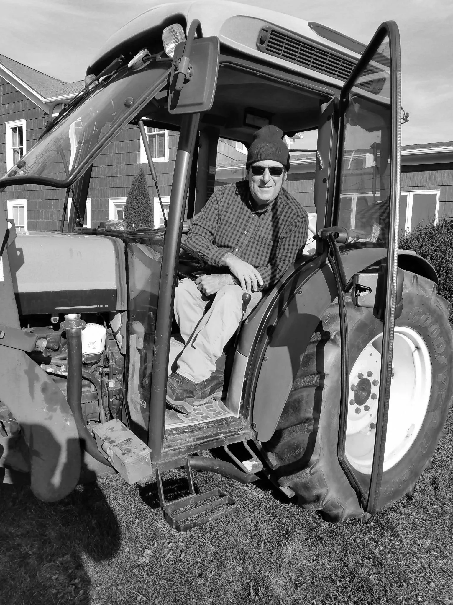 A man sitting inside a compact tractor, wearing sunglasses, a beanie, a checkered shirt, and light-colored pants, with a house and lawn in the background.