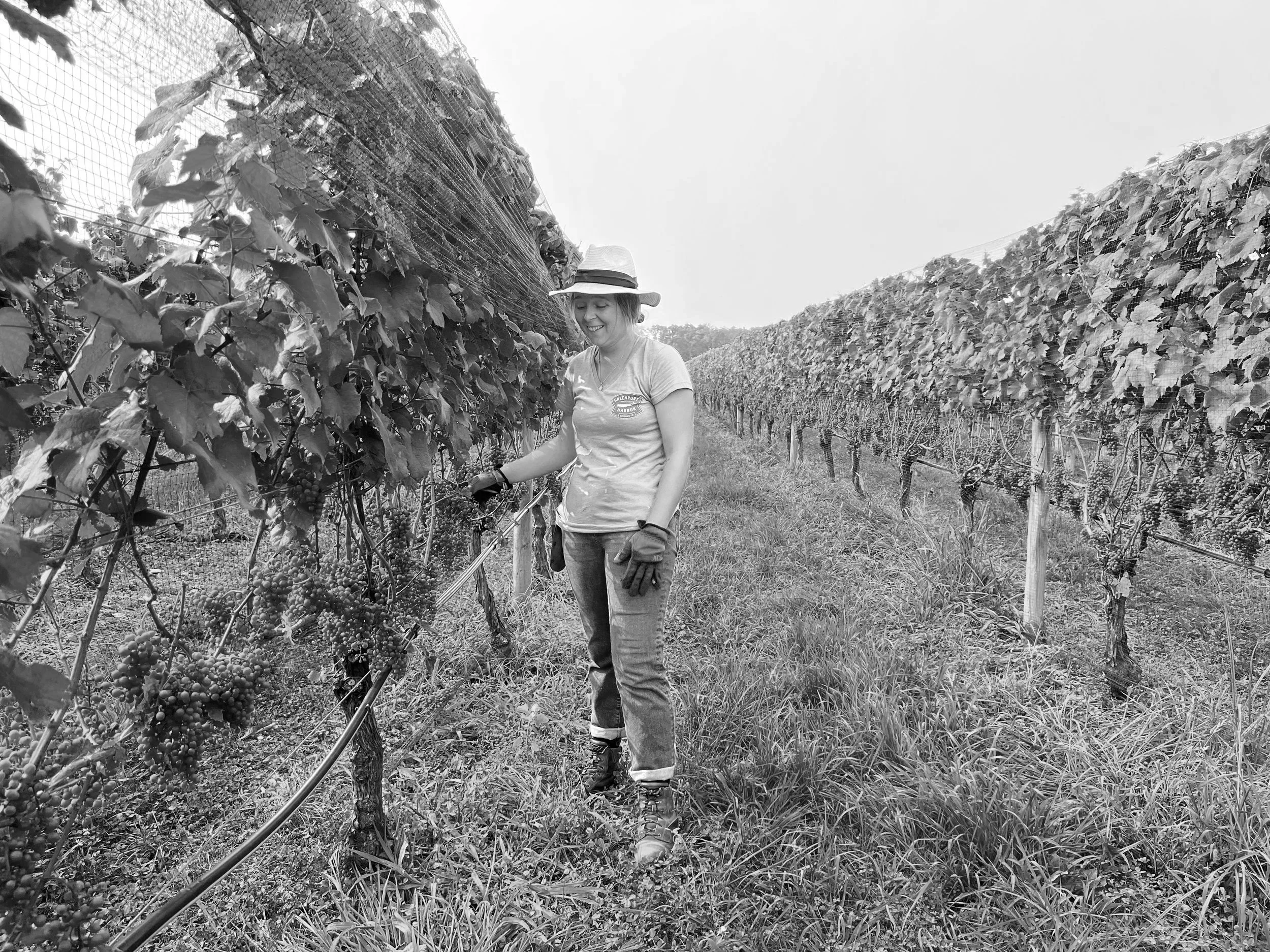 A woman smiling in a vineyard, surrounded by rows of grapevines, wearing a sun hat, t-shirt, gloves, and jeans.