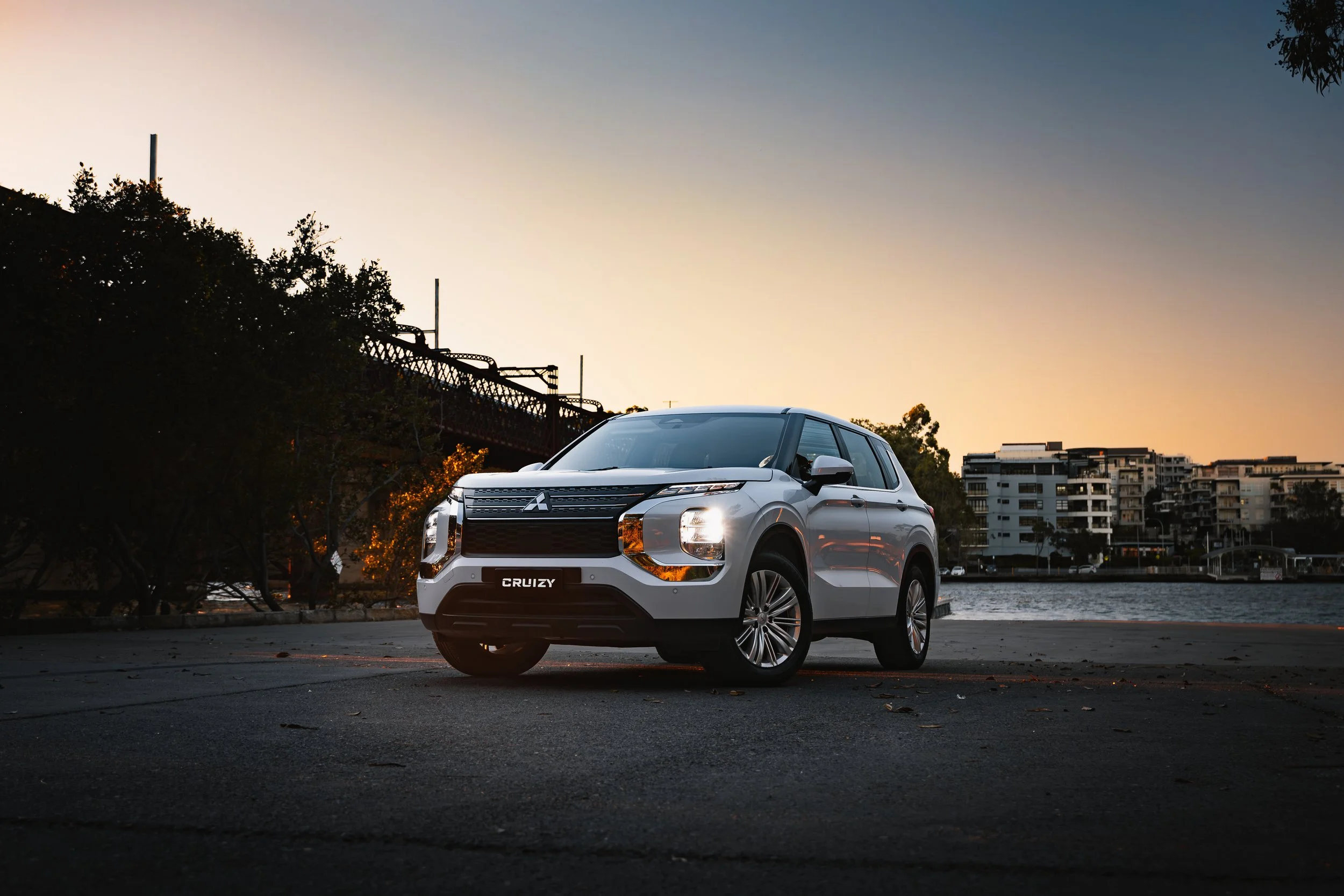A white Mitsubishi SUV parked on a paved area near water during sunset, with trees and modern buildings in the background.