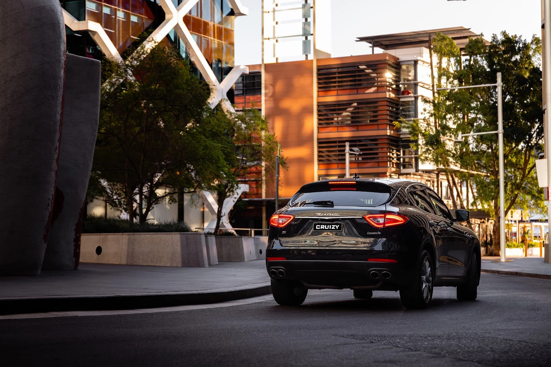 Black Maserati SUV with a license plate reading 'CRUIZY' parked on city street during sunset with modern buildings and trees in the background.