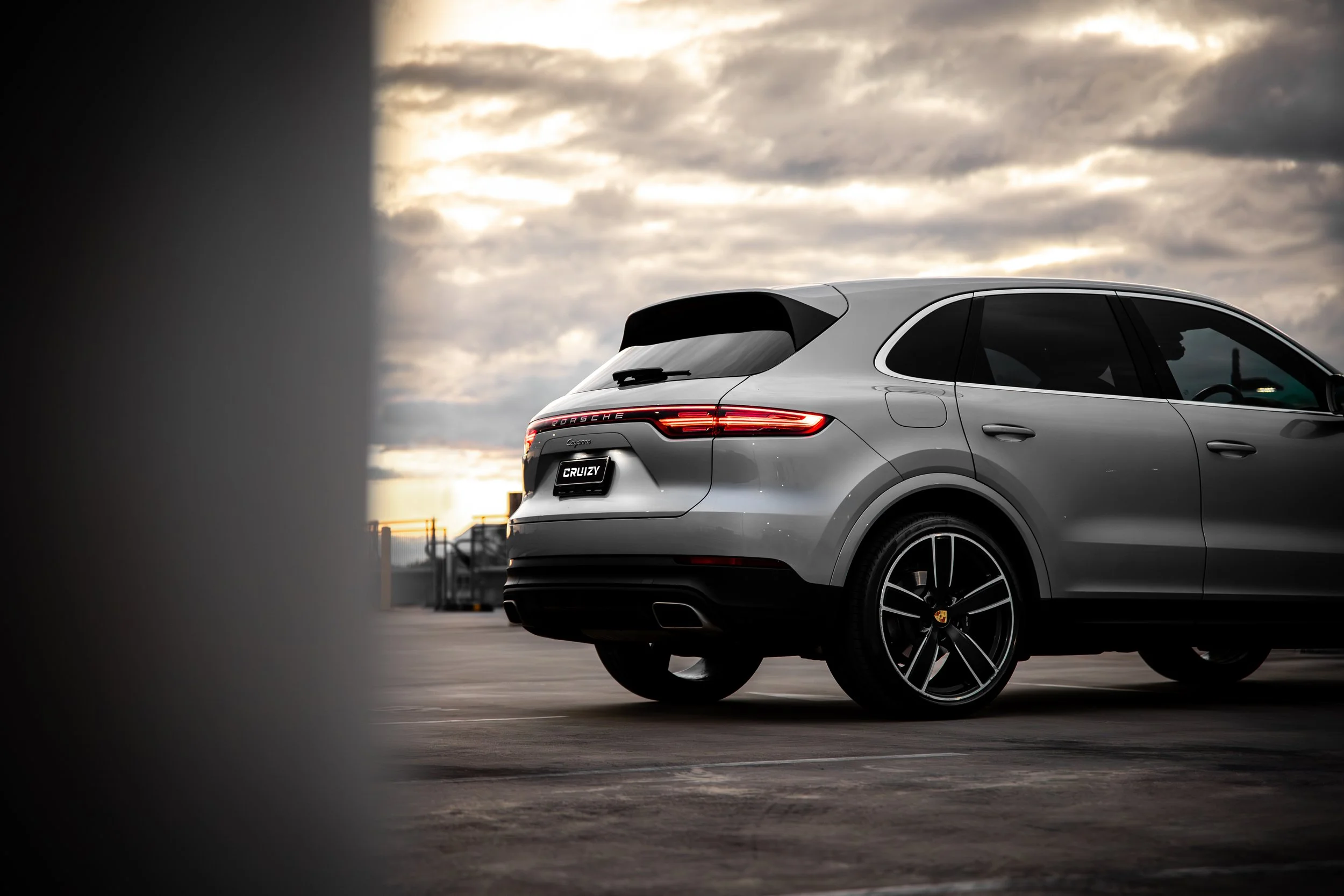 A silver Porsche SUV parked in a parking lot during sunset, with a cloudy sky in the background.