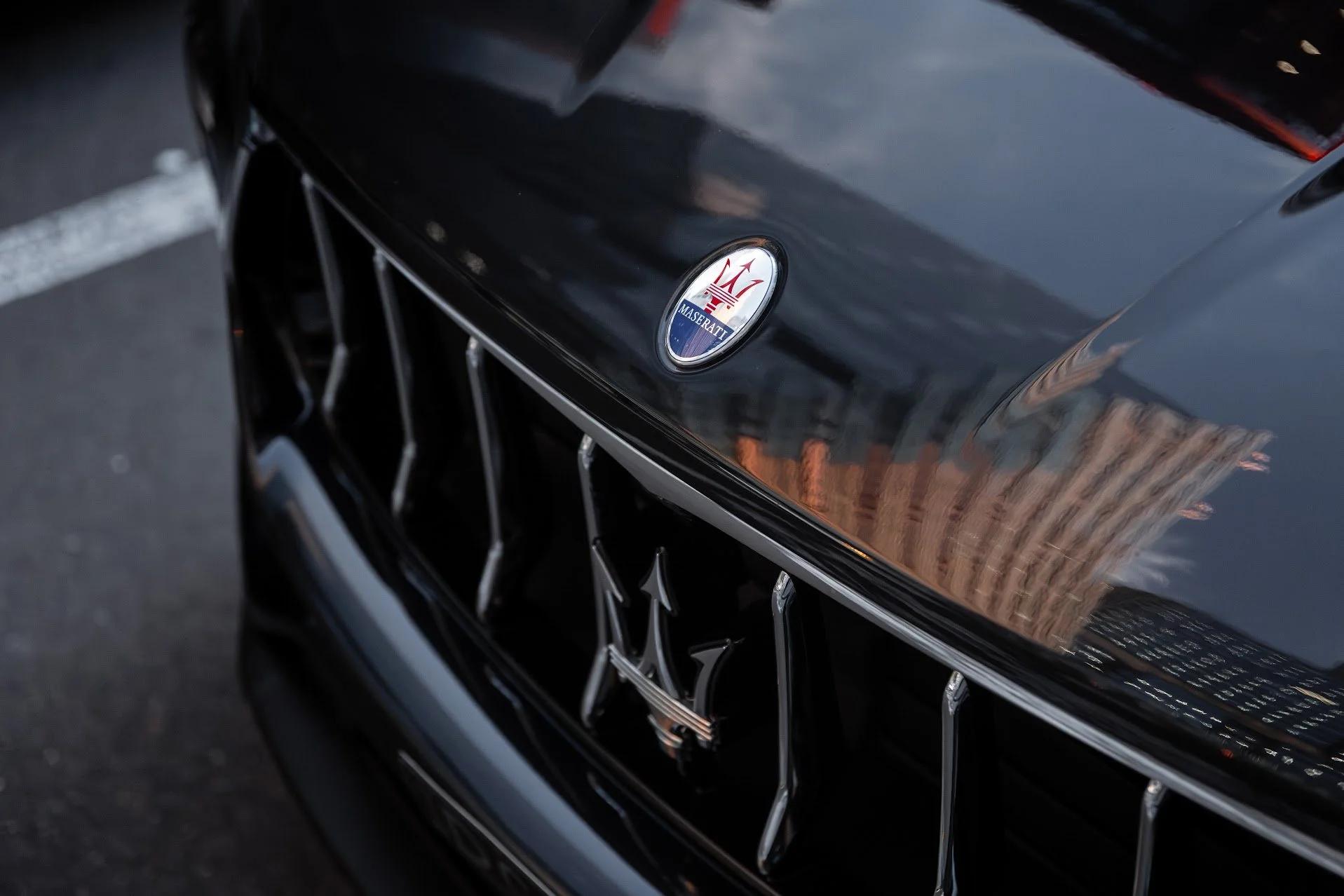 Close-up of the front grille and hood of a black Maserati car, with the Maserati emblem on the hood and reflections of buildings on the surface.