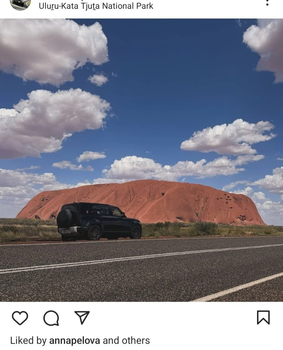 A black vehicle parked beside a road with Uluru, a large red sandstone rock formation, in the background under a partly cloudy sky.