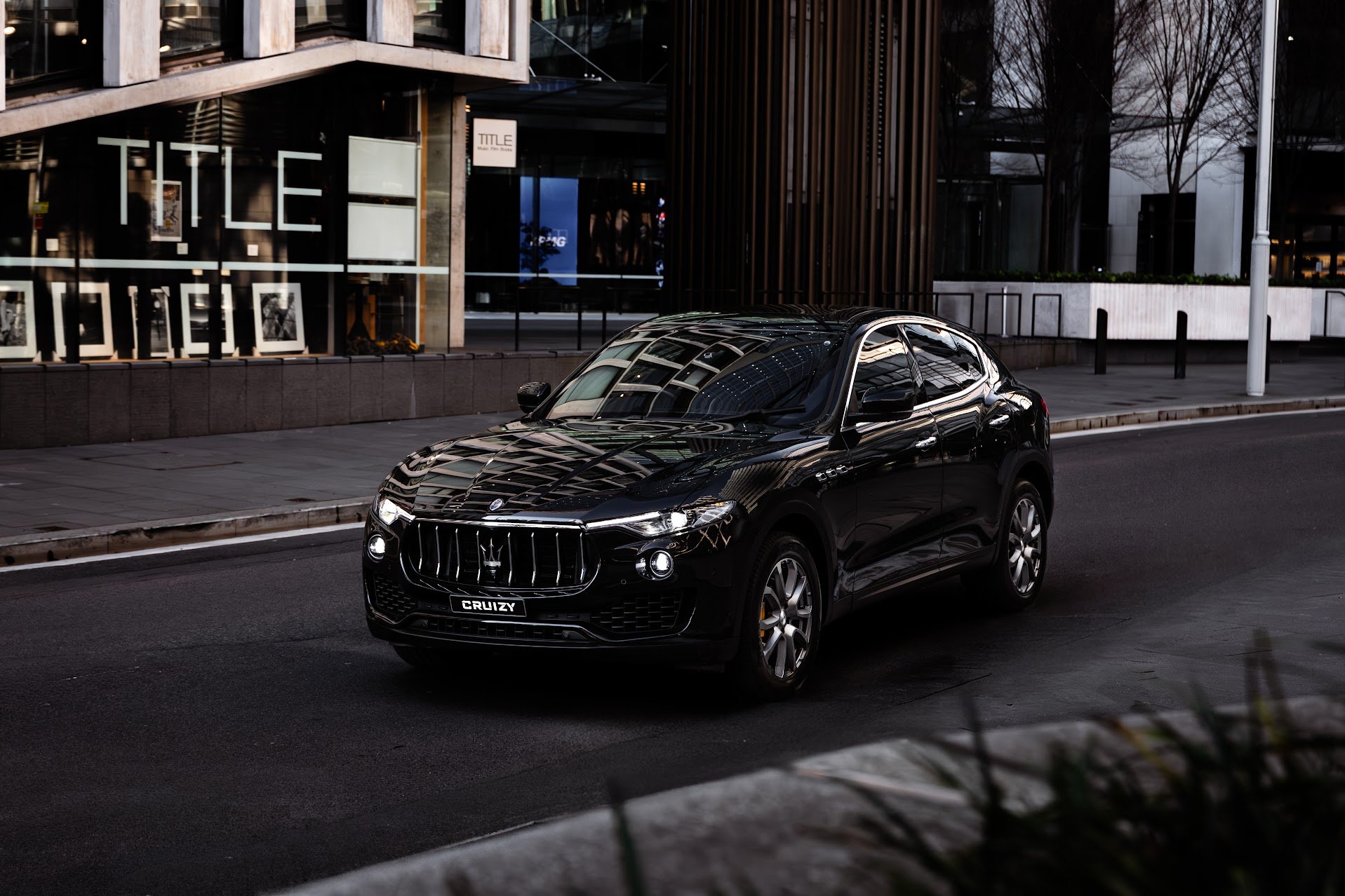 A black Maserati SUV driving on an urban street at dusk, reflecting nearby buildings on its surface.