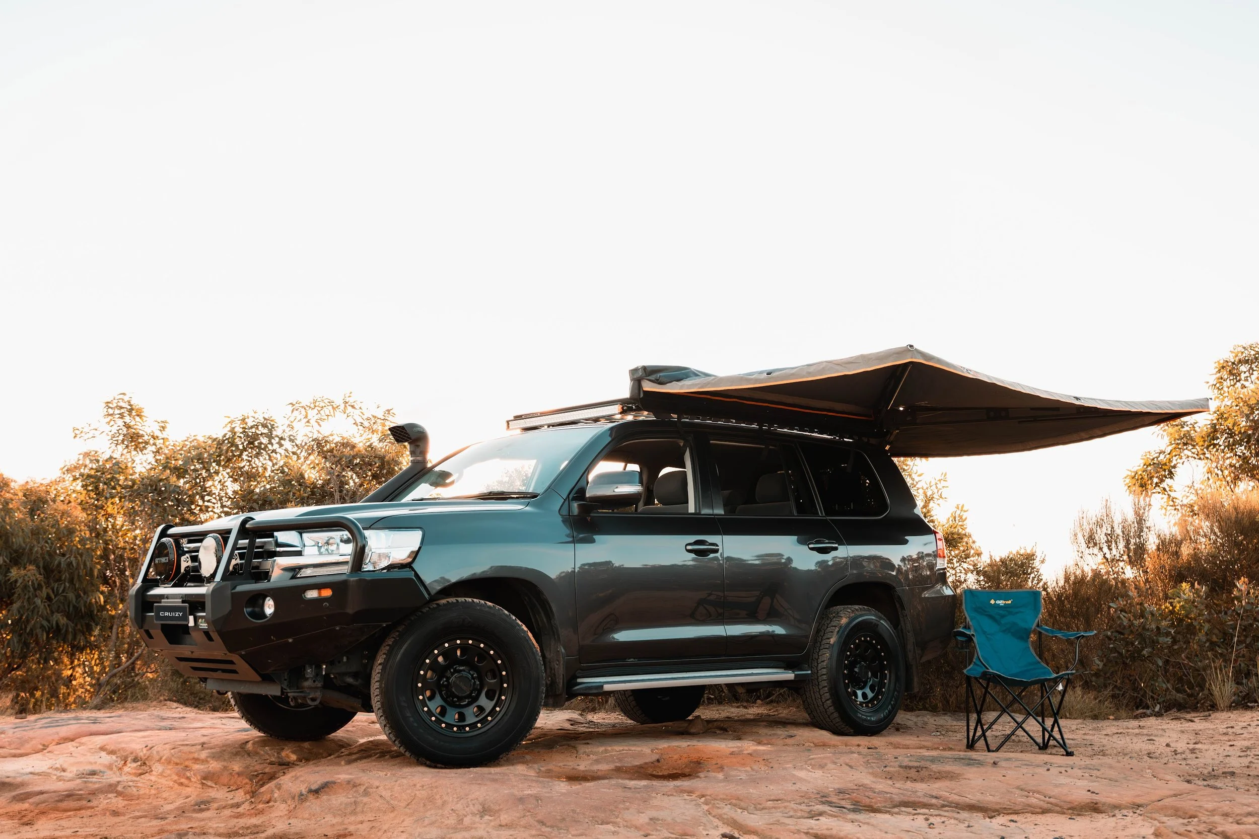 A dark gray SUV with a roof tent set up on top, parked on rocky terrain surrounded by desert vegetation and bushes during sunset.
