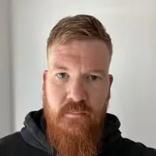 A man with a beard and mustache, short light brown hair, wearing a black hoodie, standing indoors in front of a light-colored wall.