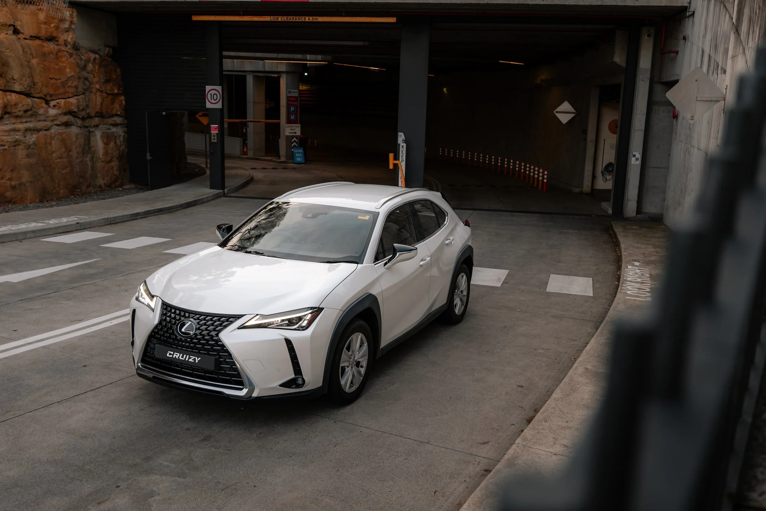 A white Lexus UX hybrid car parked near an underground parking entrance, with signs indicating speed limit and parking regulations, in a dimly lit urban setting.