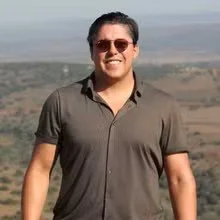 Man smiling outdoors with a landscape background.