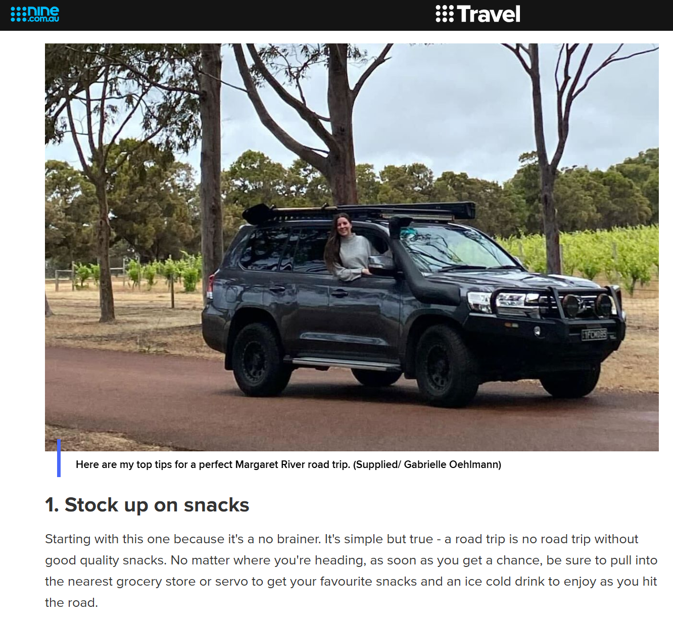 A woman sitting out of a black SUV parked on a dirt road with trees and vineyard in the background.