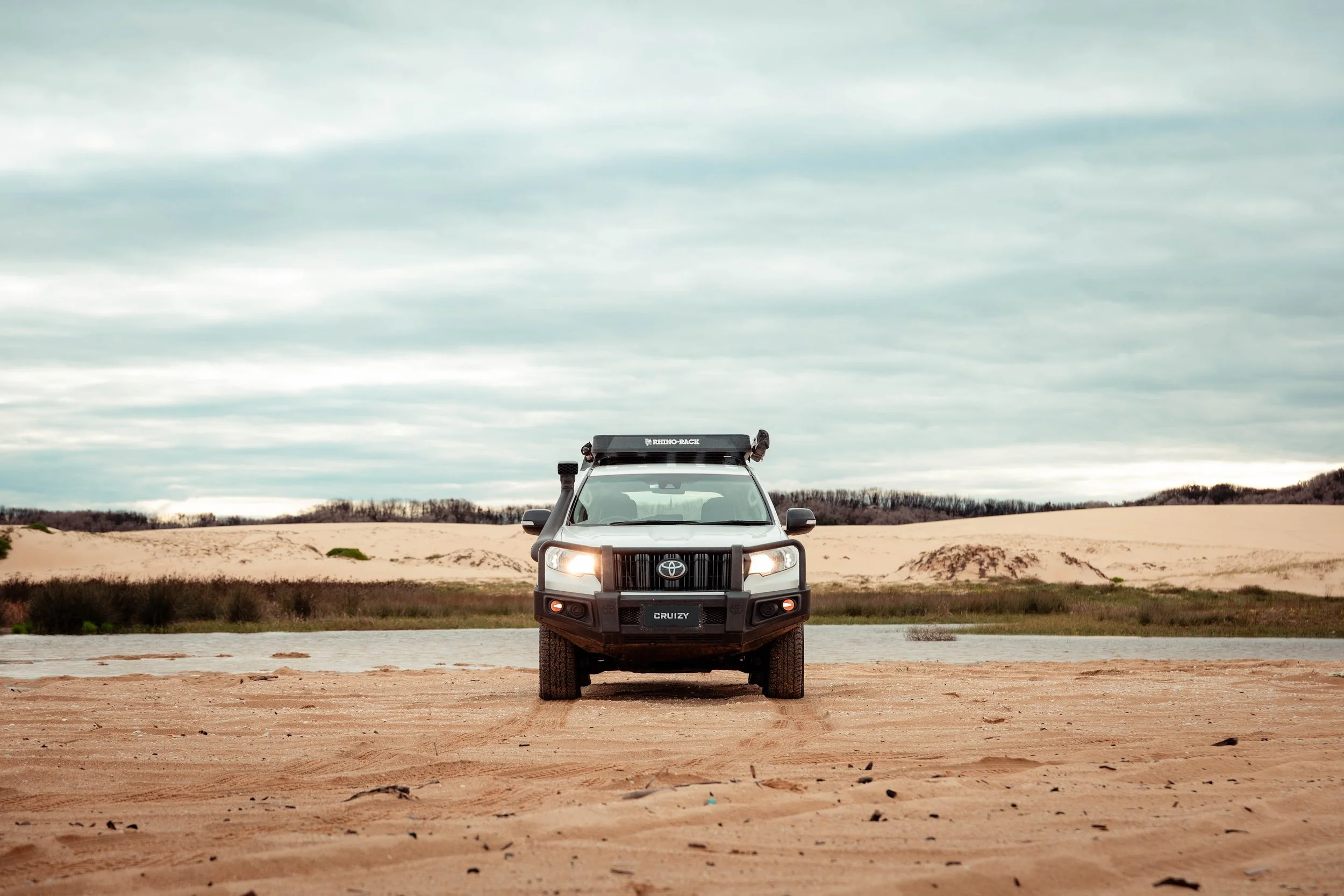 Black Toyota Cruiser with rooftop tent parked on sandy terrain, with a water body, grassy area, and sand dunes in the background under a cloudy sky.