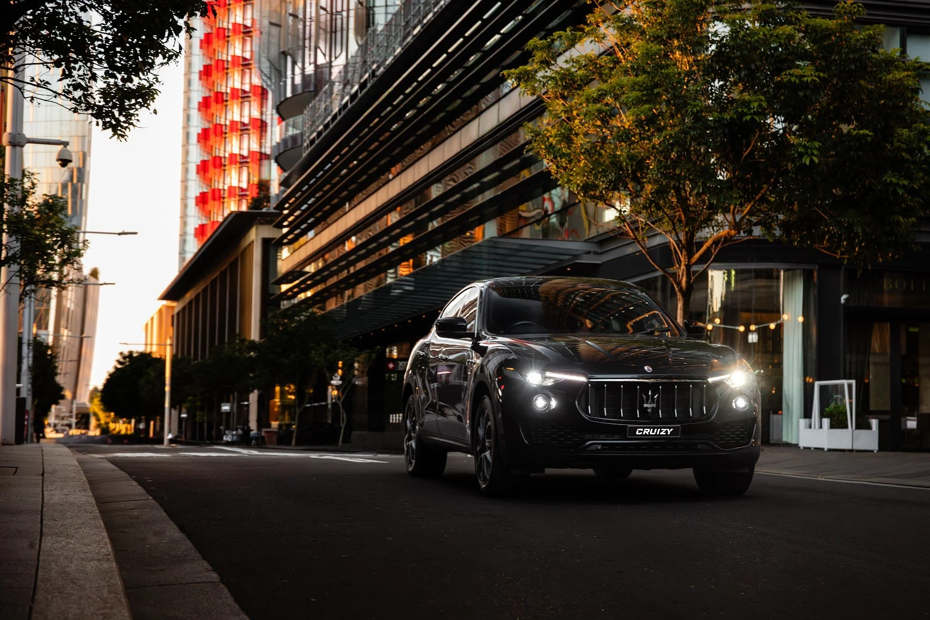A black Maserati SUV driving on an empty city street during dusk, with modern buildings and trees lining the sidewalk.