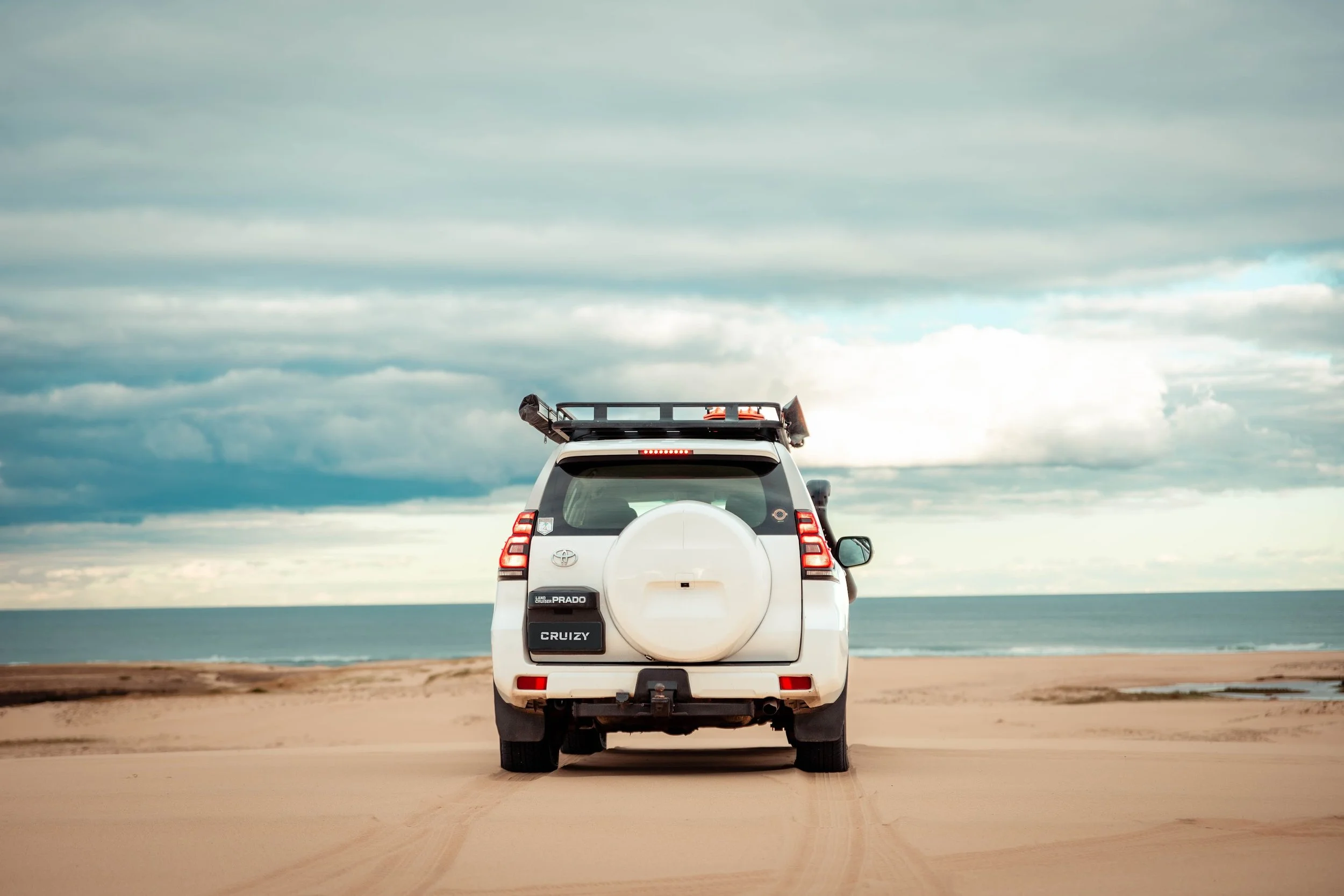 White Toyota Land Cruiser Prado parked on sandy beach facing the ocean, with a cloudy sky overhead.
