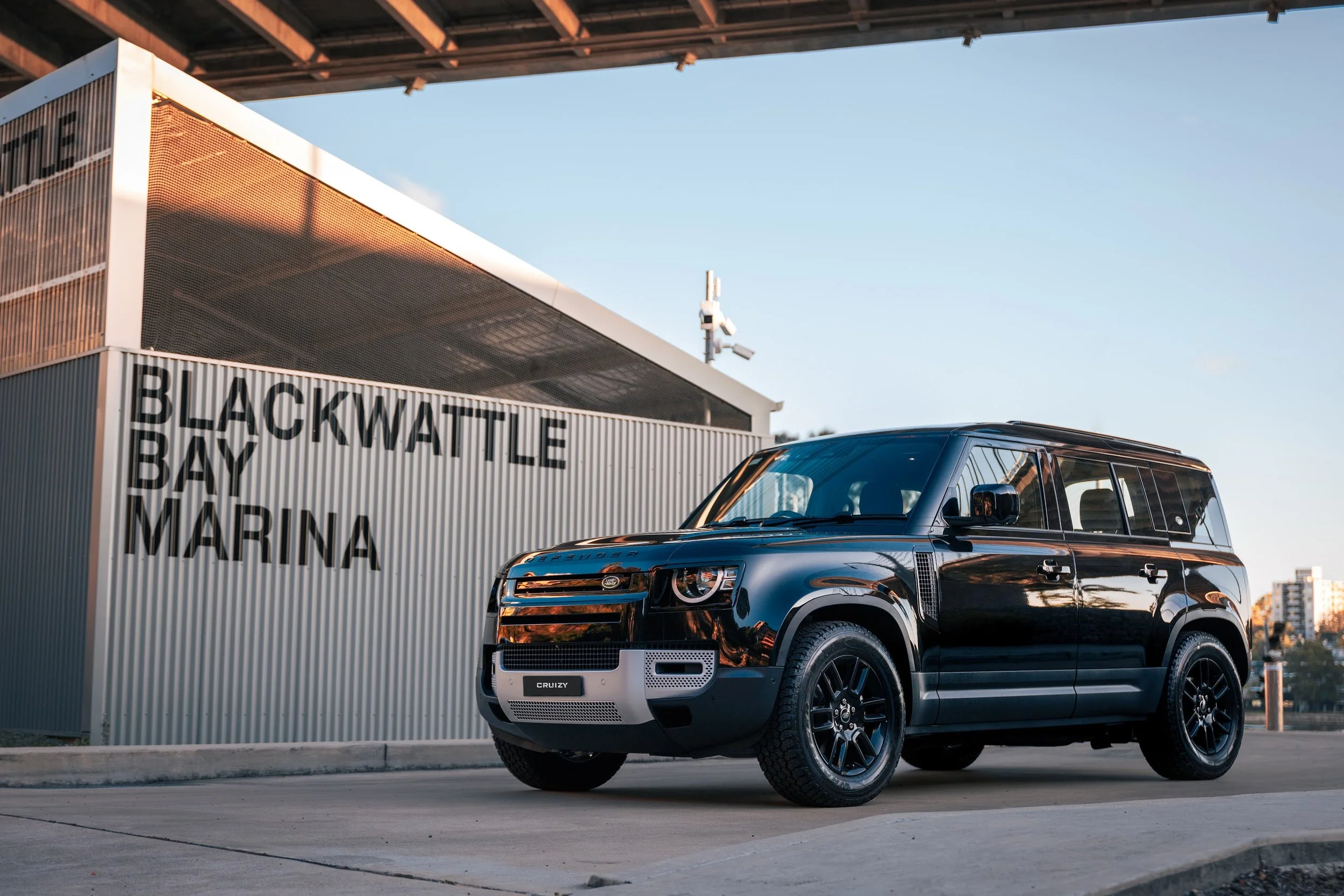 A black Land Rover Defender parked in front of a building at Blackwattle Bay Marina during sunset, with city buildings visible in the background.