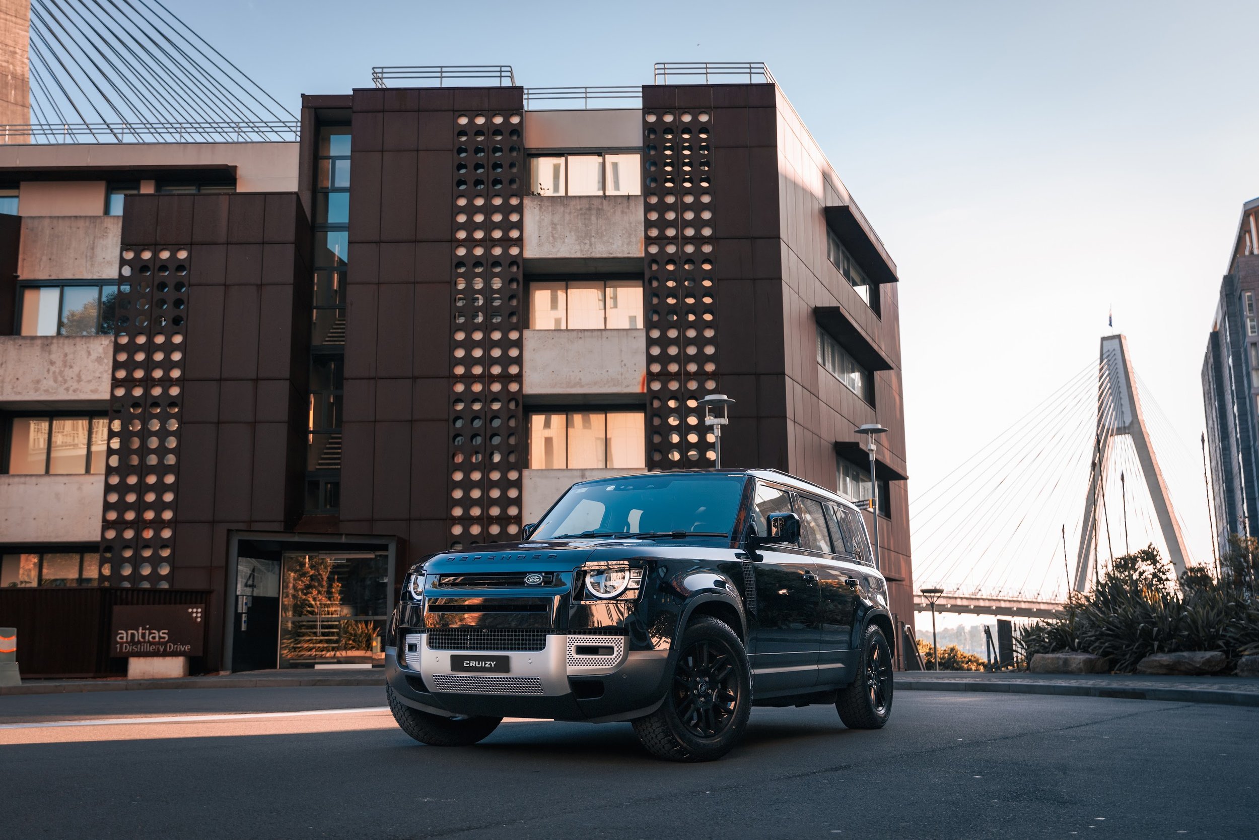 A black Land Rover Defender SUV parked on city street in front of modern building with perforated facade, and a bridge with cables in the background during sunset.