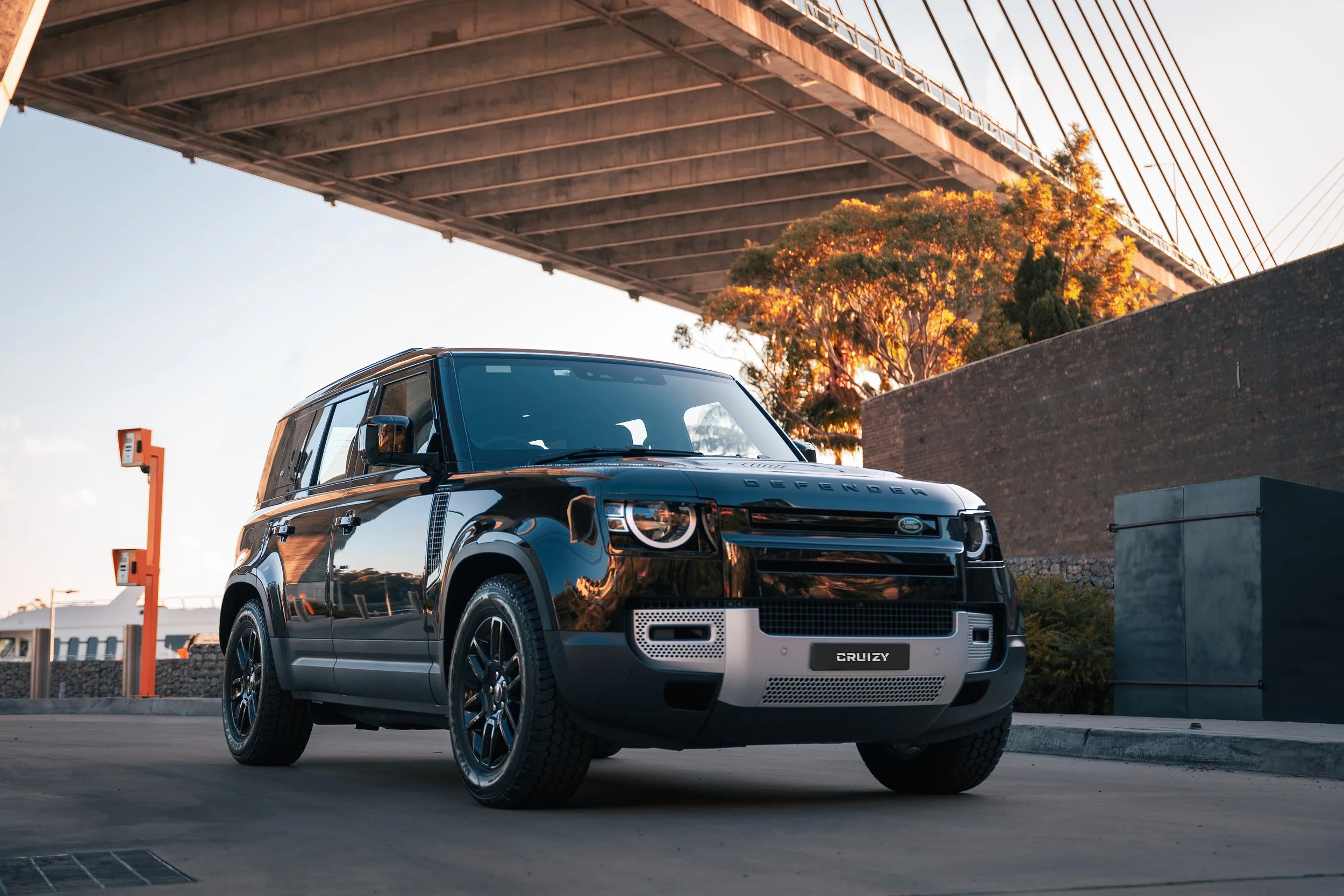 Black Land Rover Defender Cruzy parked under a bridge during sunset with trees and a brick wall in the background.