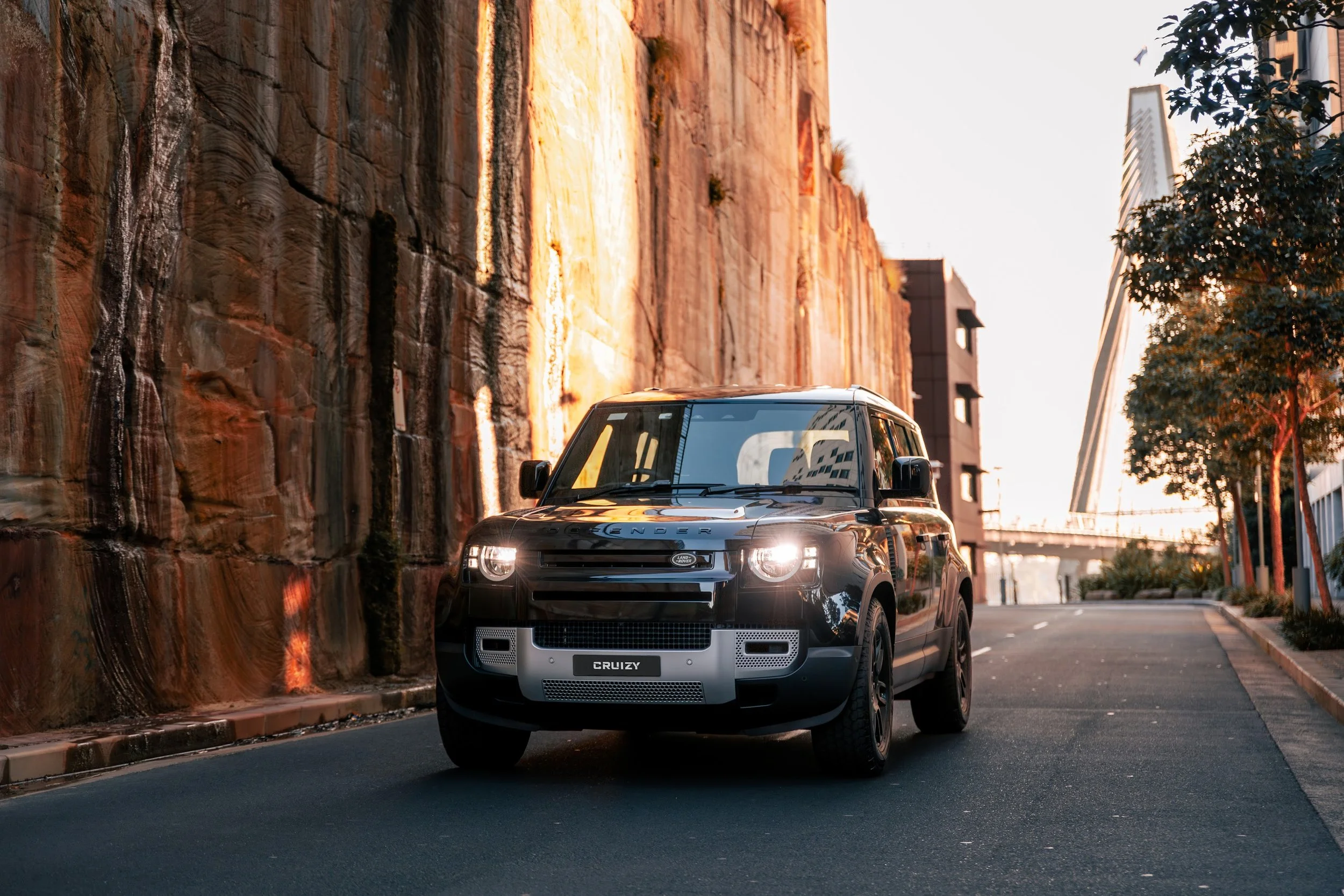 A black Land Rover SUV driving on an urban street at sunset, with a large rock wall on one side and modern buildings and trees in the background.