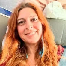 Smile of a woman with long red hair, wearing a colorful shirt, indoors with a blurred background.