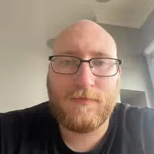 A young man with glasses and a beard taking a selfie indoors, with a light-colored wall and window in the background.