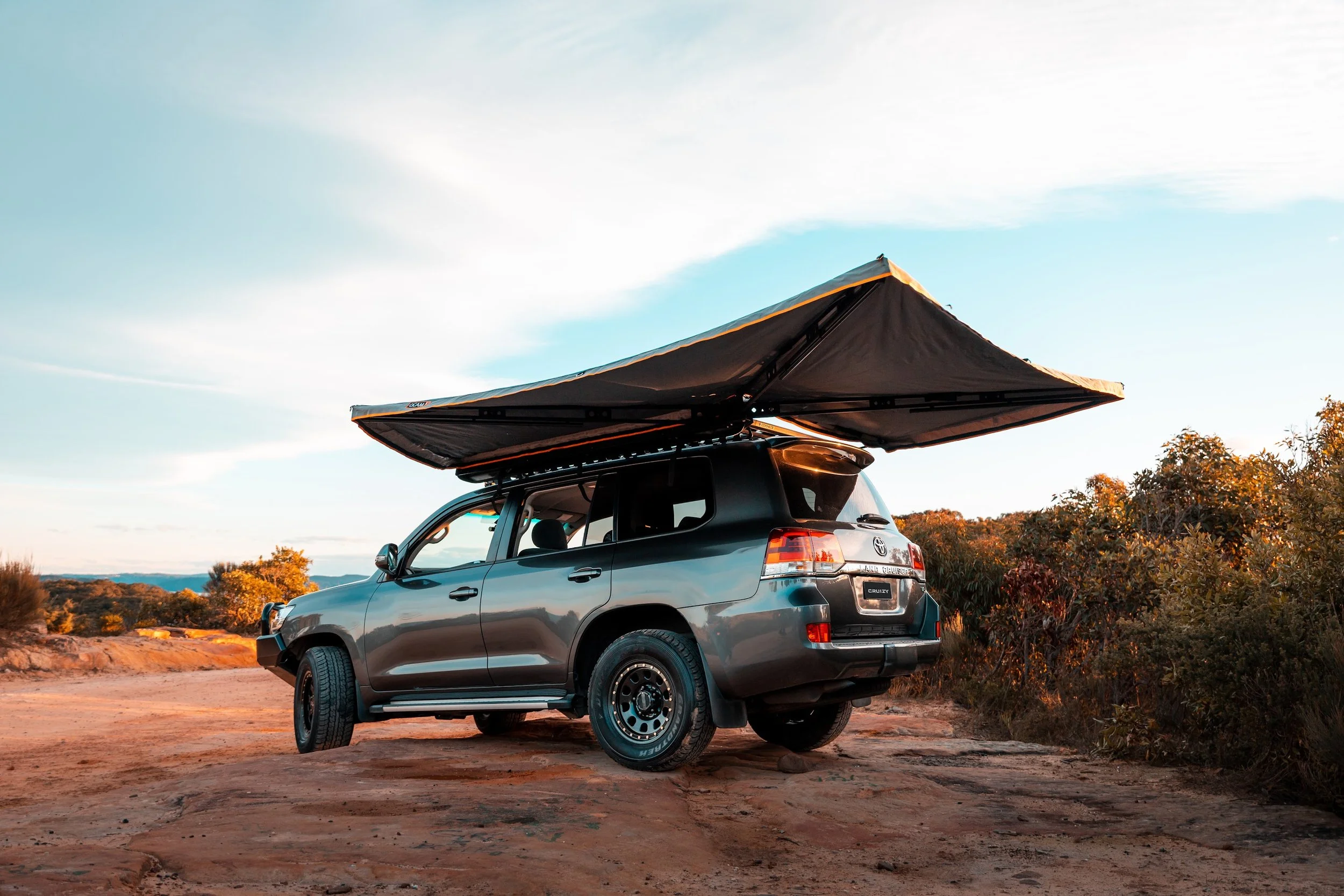 A gray SUV parked on a dirt surface with a rooftop awning extended, capturing a scenic outdoor landscape at sunset.