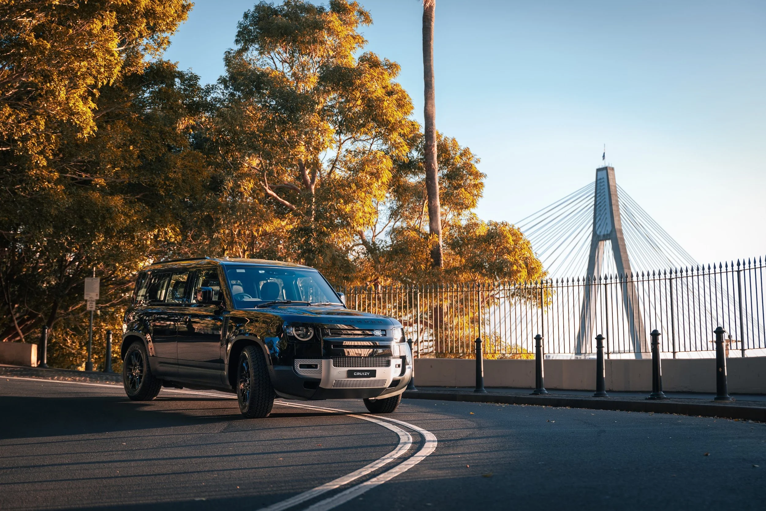 A black SUV parked on a curved road with a double white line, gold-leaved trees and a modern cable-stayed bridge in the background during sunset.