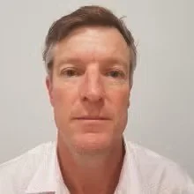 Close-up photo of a man with short brown hair wearing a white shirt, against a plain light background.