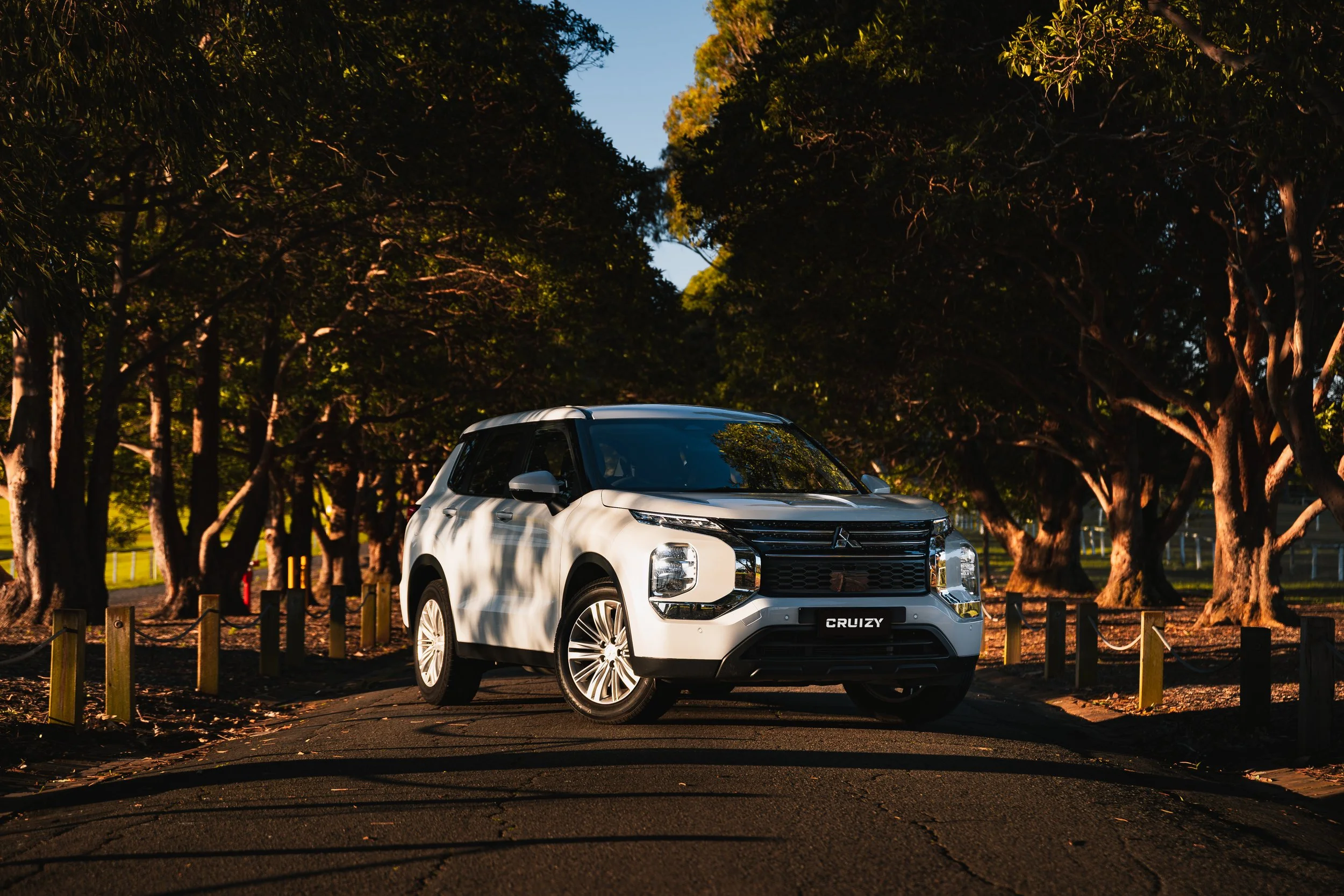 A white Mitsubishi SUV parked on a paved road surrounded by trees with green leaves, with sunlight filtering through the branches, creating shadows.