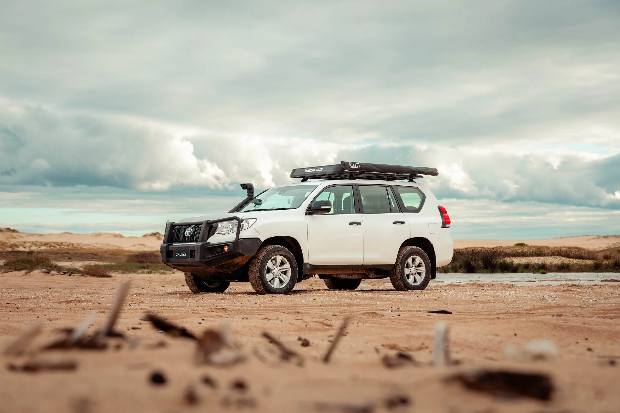 White Toyota SUV with roof rack and surfboard driving in a desert landscape under cloudy sky.