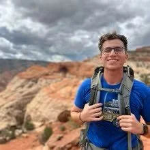 A young man with glasses wearing a blue shirt and backpack, smiling outdoors with a red rock canyon and cloudy sky in the background.