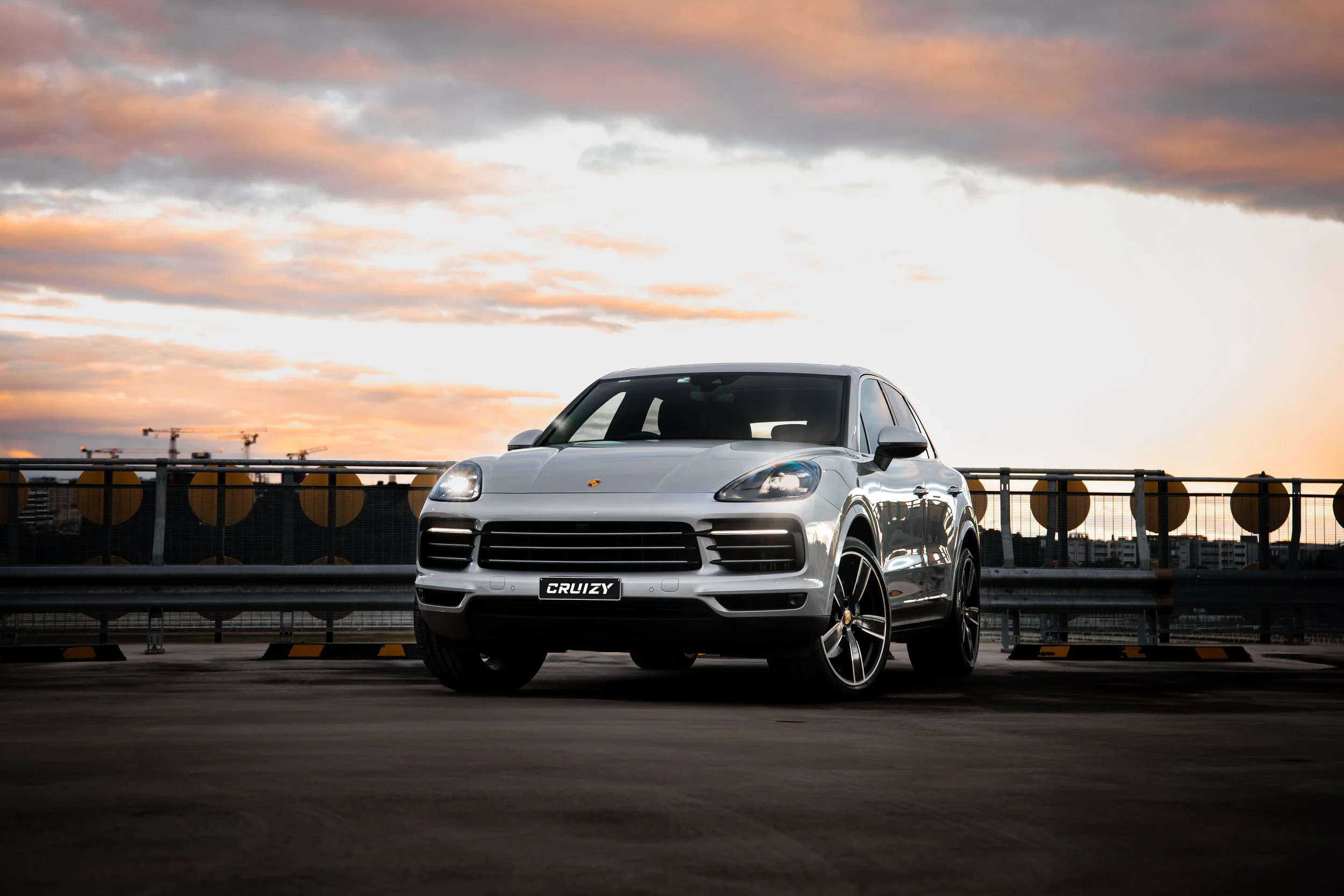 A white Porsche SUV parked on a rooftop parking lot at sunset, with a city skyline and construction cranes in the background.