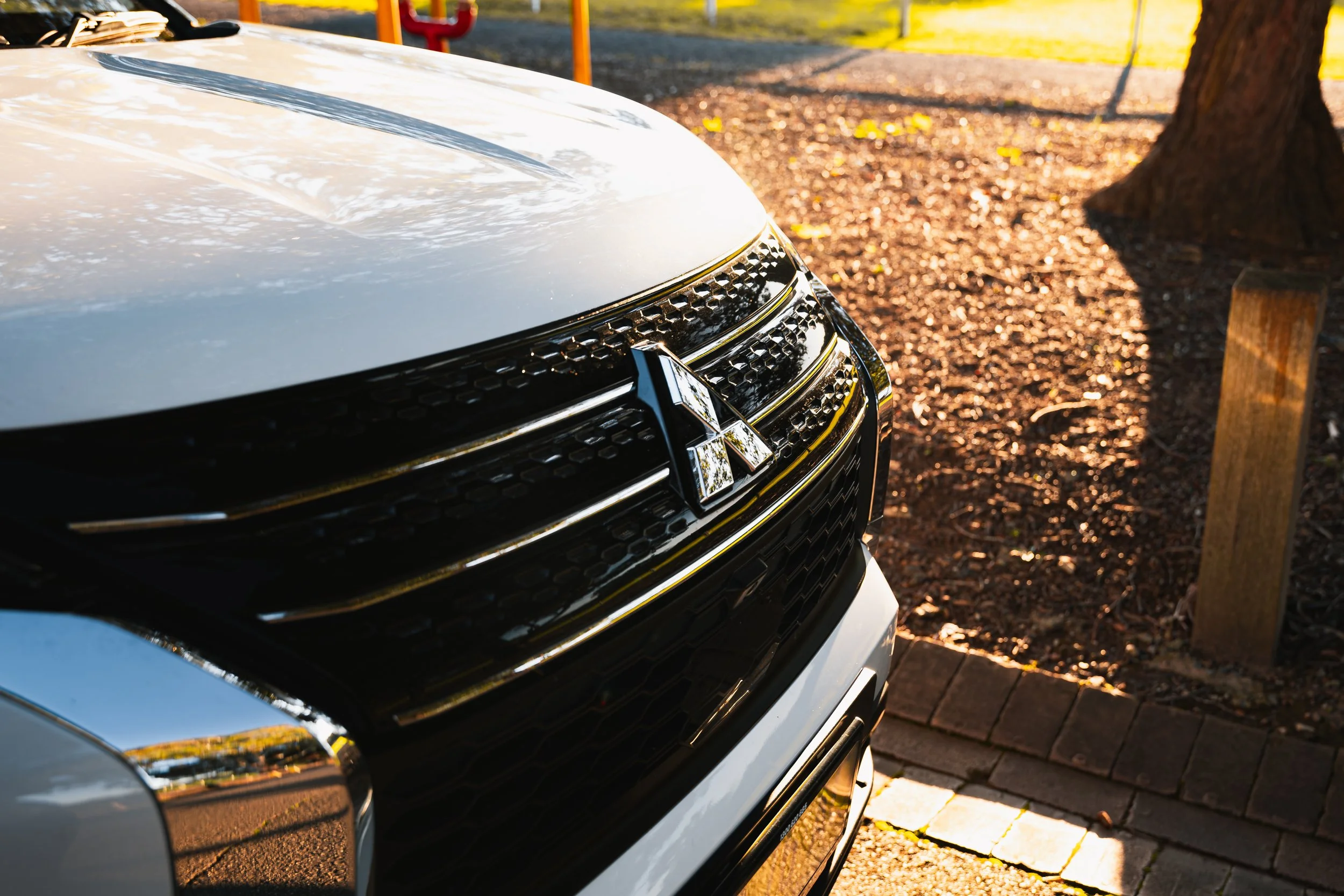 Close-up of the front of a white Mitsubishi vehicle, showing the black grille and chrome emblem, parked on a brick-paved area near a tree and a wooden post, with warm sunlight reflecting on the car.