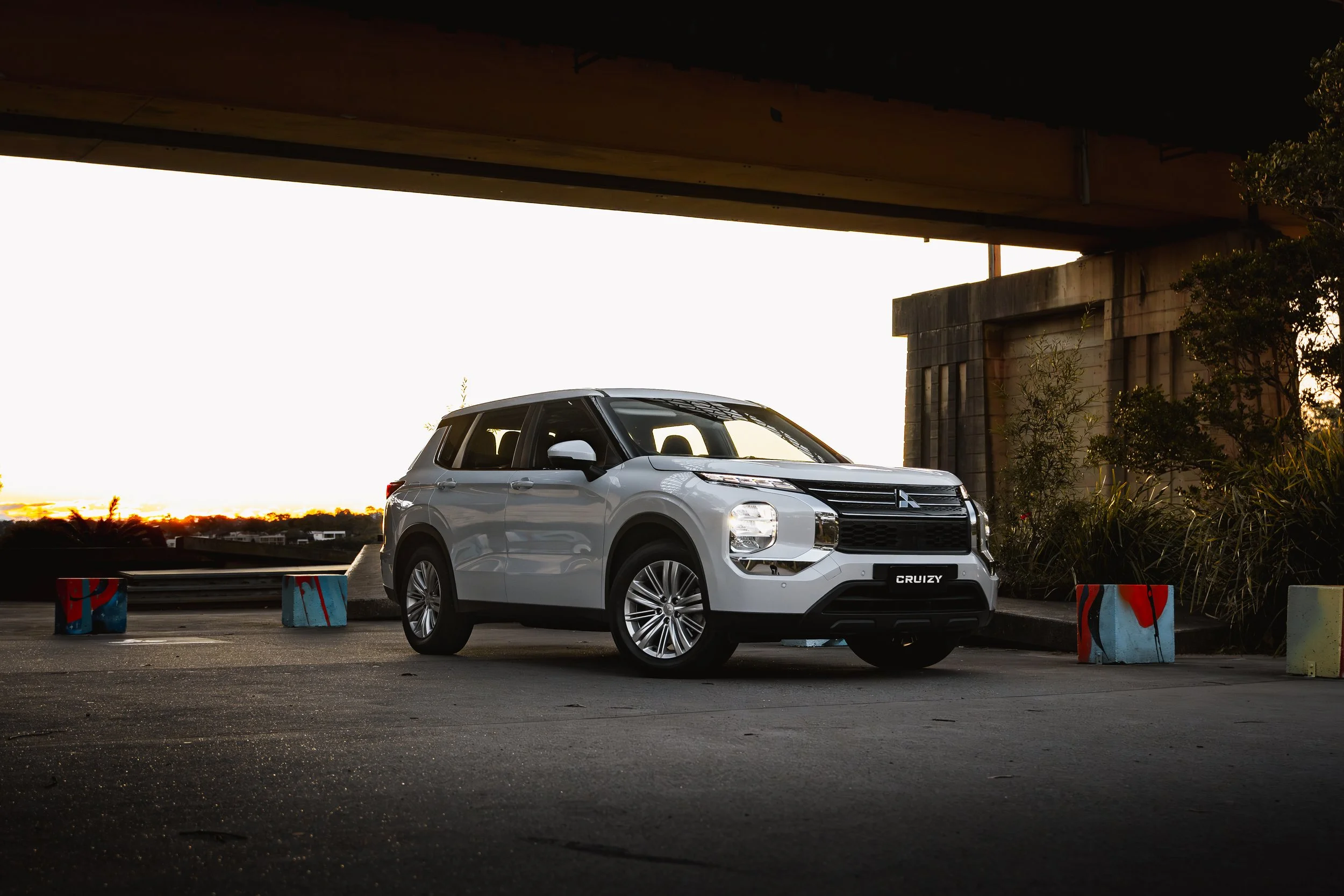 A silver Mitsubishi SUV parked under a bridge at sunset.
