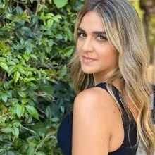 Young woman with long wavy blonde hair wearing a black top, standing outdoors near green foliage, looking at the camera with a slight smile.