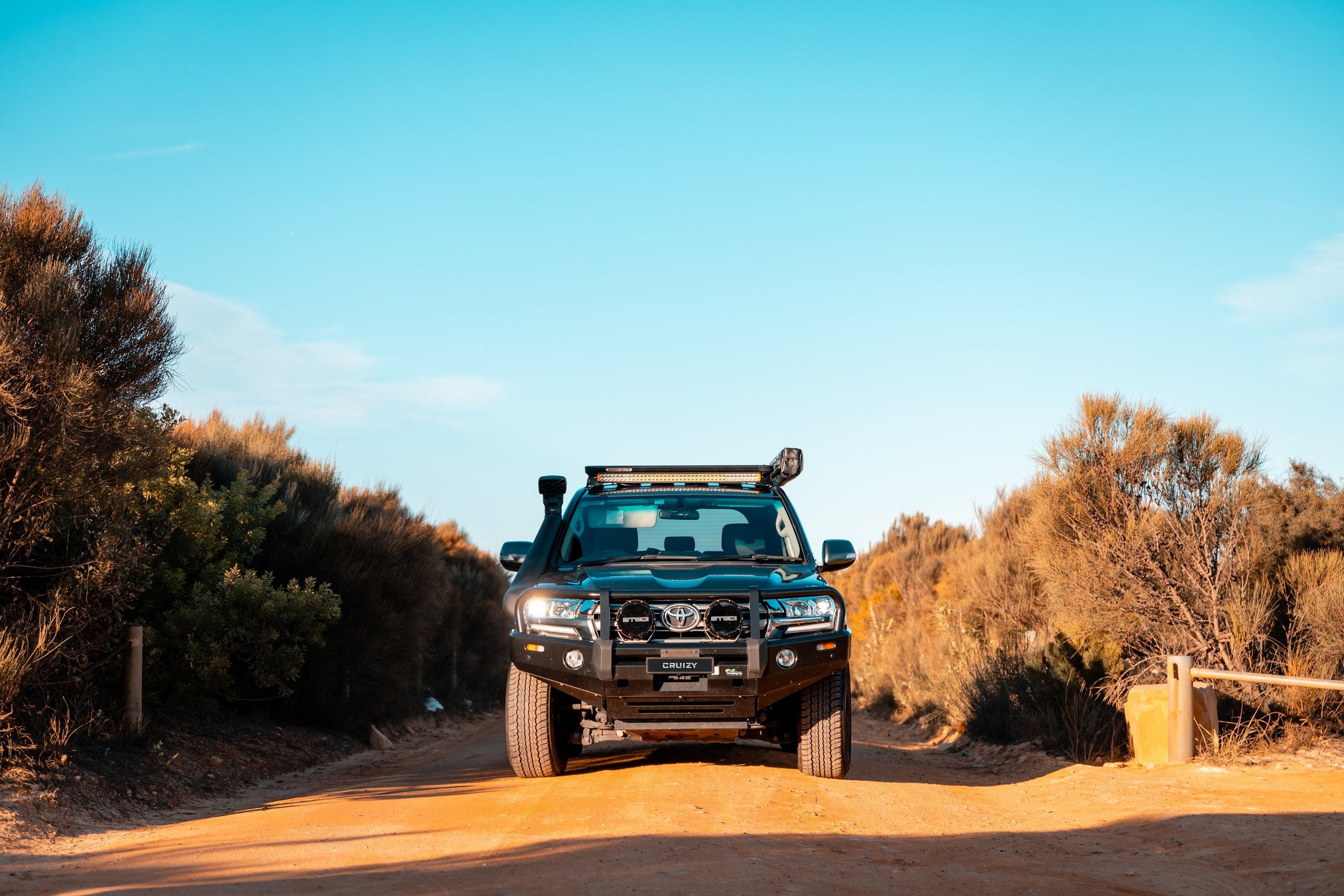 A black off-road vehicle with a light bar and auxiliary lights parked on a sandy dirt road surrounded by desert shrubs and trees, under a clear blue sky.