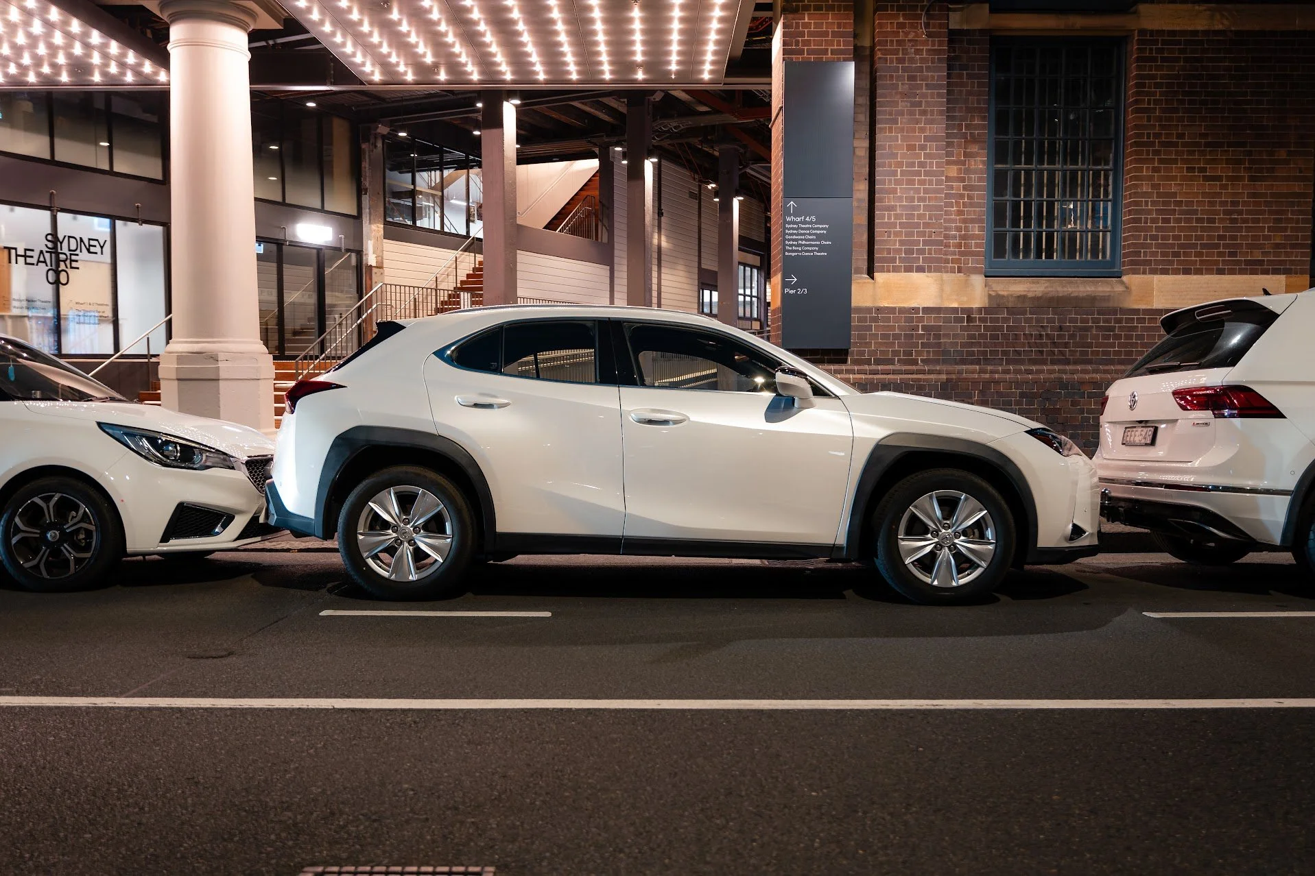 White hatchback and crossover parked on a city street at night, with brick building and signage in the background.