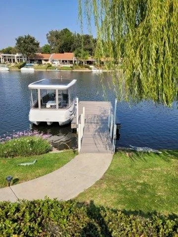 A dock with a boat moored to it, extending into a calm body of water near residential houses with lush trees, under a clear blue sky.