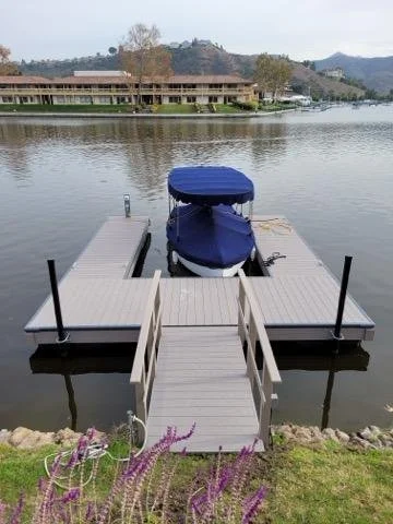 A boat covered with a blue tarp docked at a wooden pier on a calm lake with houses and hills in the background.