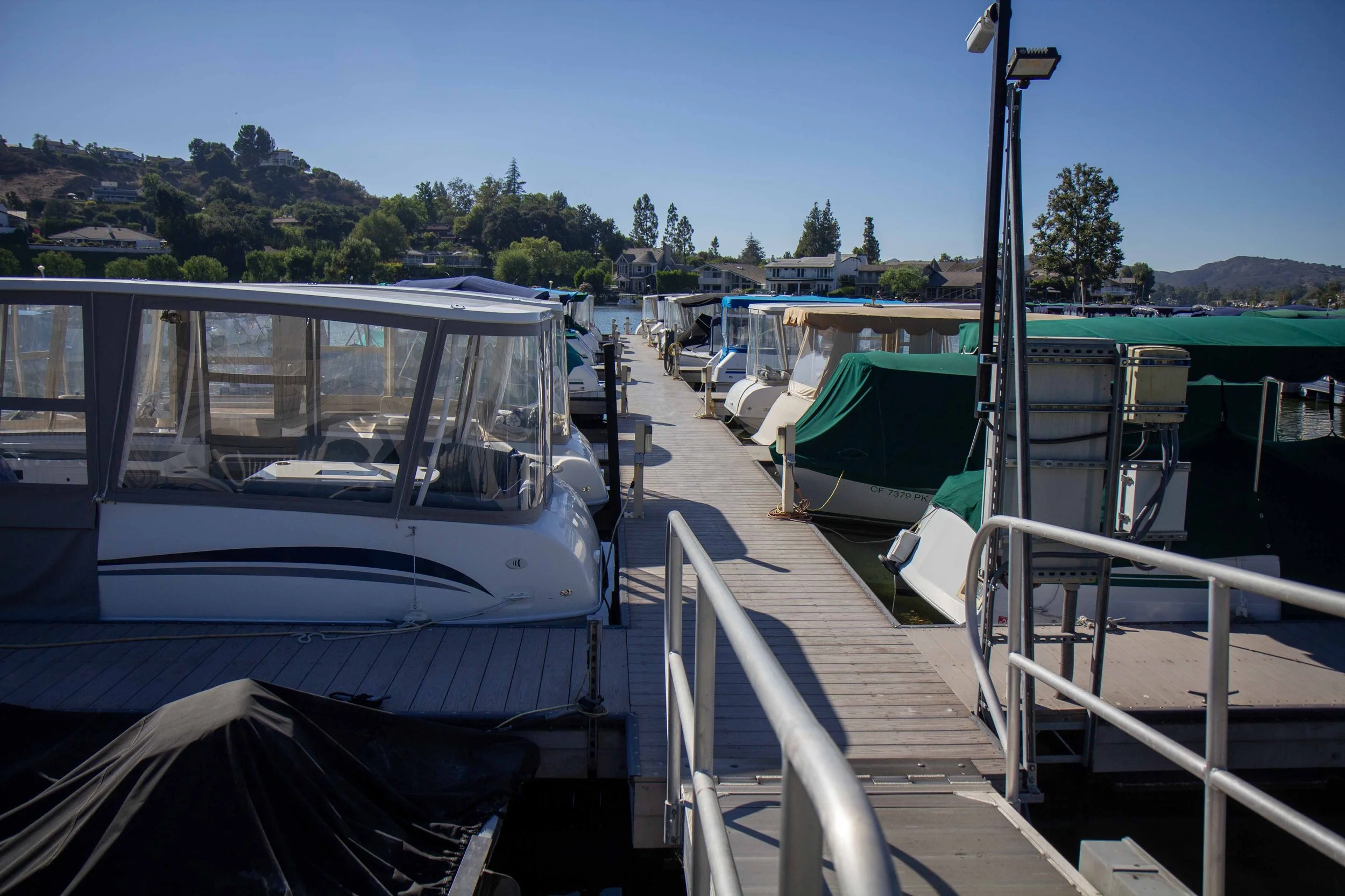 Boats docked at a marina on a sunny day, with houses and trees on a hillside in the background.